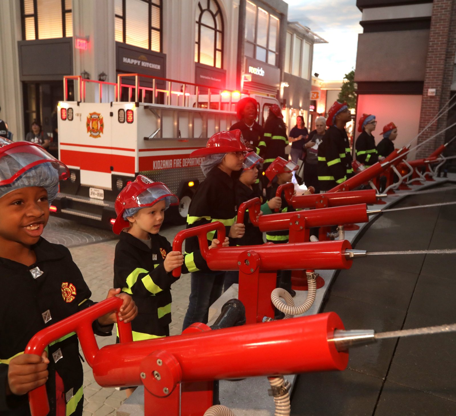 Children in firefighter gear using water cannons next to the KidZania fire truck