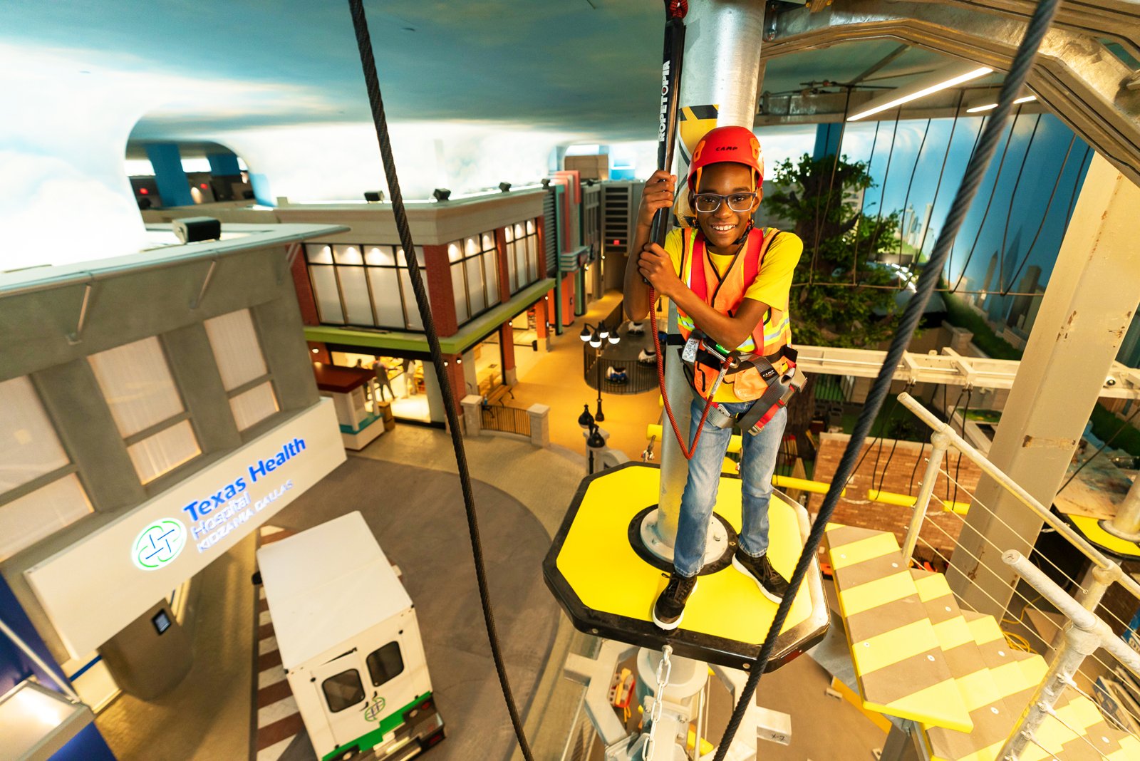 Child in a harness and helmet on a high ropes platform above the KidZania city